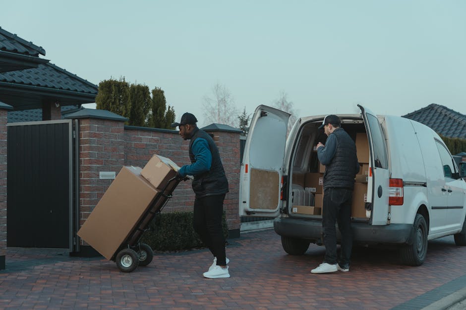 Two delivery workers are unloading boxes from a white van parked on a brick driveway in a residential area with houses and trees in the background. One worker is pushing a cart loaded with large cardb