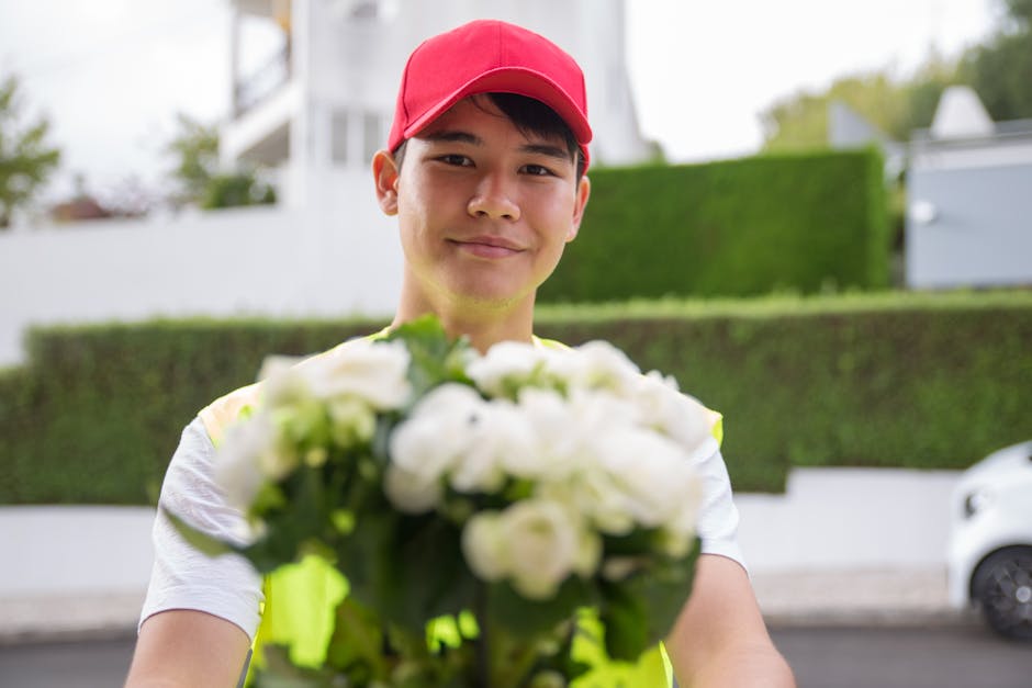 A delivery person wearing a red cap and a high-visibility vest is smiling warmly while extending a bouquet of fresh white flowers towards the camera. The bouquet, held in their hands, features large, 