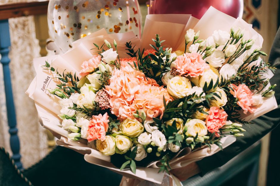 A vibrant floral arrangement featuring a variety of fresh flowers, including soft pink carnations, white roses, and peach-colored blooms, wrapped in elegant cream-colored tissue paper with a subtle sh