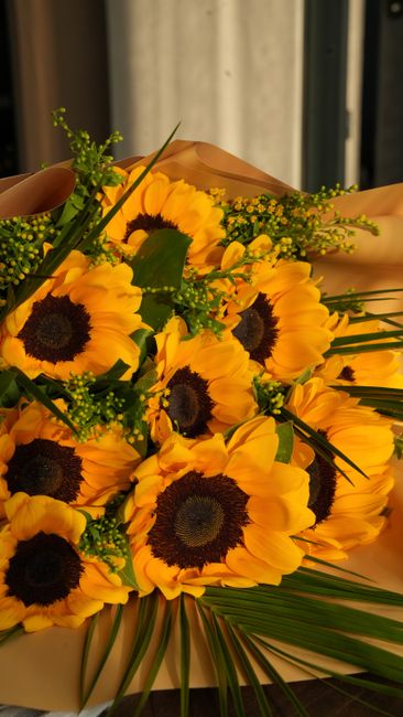A vibrant bouquet of fresh sunflowers with large yellow petals and dark brown centers, arranged with green foliage and small yellow filler flowers. The bouquet is wrapped in light brown craft paper an