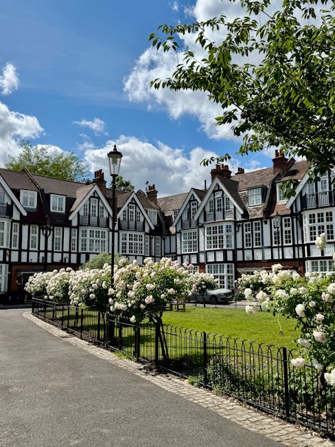 A row of traditional UK-style houses with white walls and dark timber framing, set against a bright blue sky with scattered clouds. In the foreground, a neatly maintained garden features blooming whit