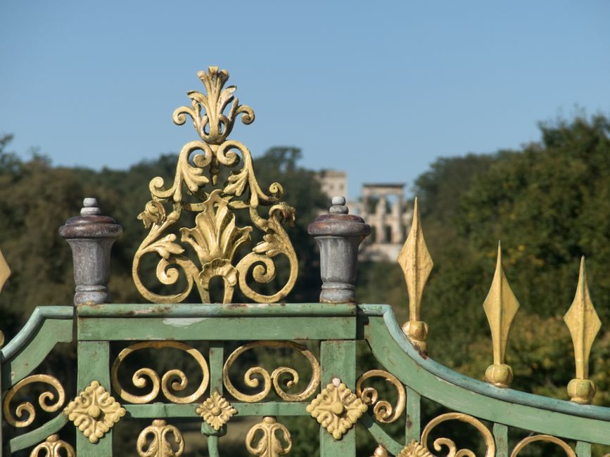 An ornate wrought iron garden gate painted in faded green with decorative gold accents, featuring intricate scrollwork and floral motifs. The top of the gate displays a central heraldic crest with scr