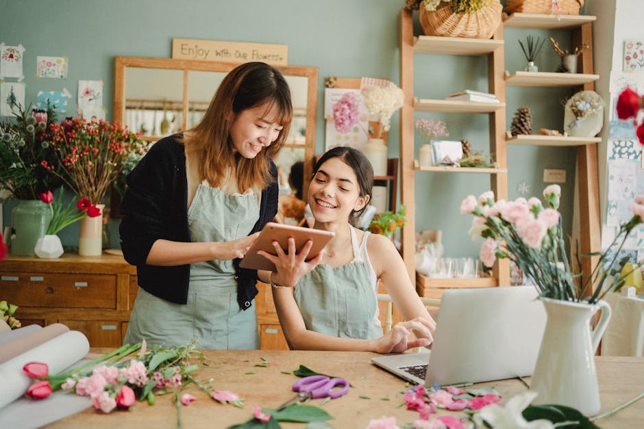 Two women working together in a bright flower shop, surrounded by vibrant fresh flowers including pink carnations and red tulips. One woman has shoulder-length brown hair and is wearing a black cardig