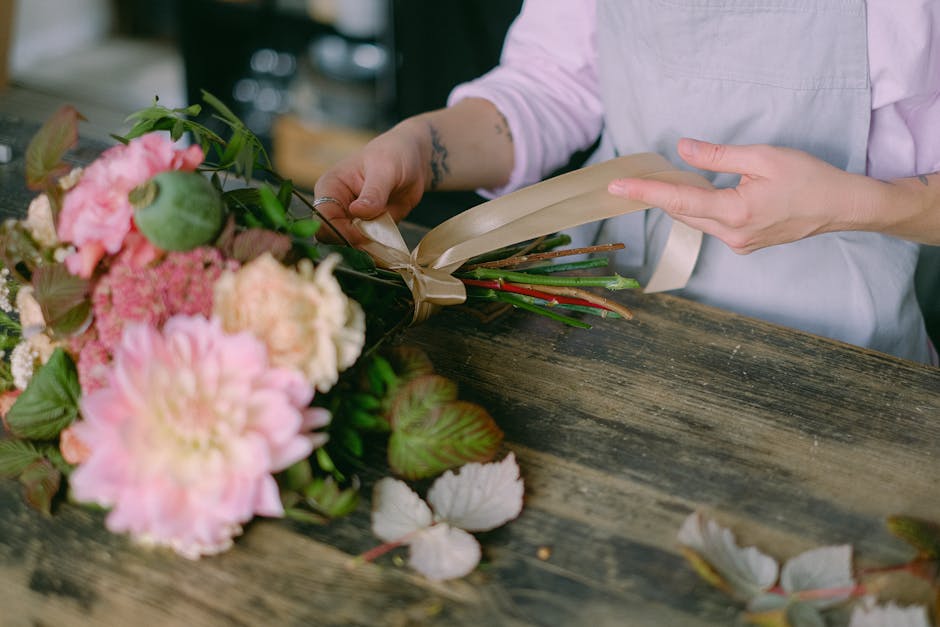 A florist's hands are shown wrapping a mixed bouquet of fresh flowers, including pink peonies, purple hydrangeas, and green foliage, with beige floral wrapping paper and a cream satin ribbon. The bouq