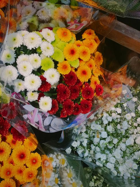 A vibrant bouquet of fresh chrysanthemums and daisies arranged in a rounded cluster, featuring bright white, orange, yellow, and red blooms. The flowers are wrapped in clear cellophane with colorful r