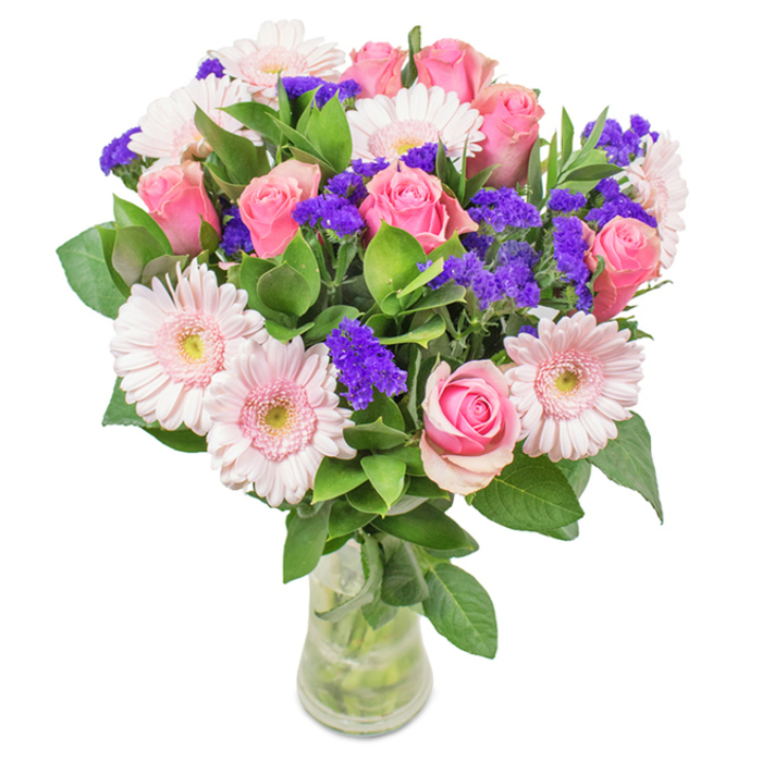 A strikingly elegant floral arrangement rests in a clear glass vase, set against a pristine white canvas, embodying the vibrant character of Dollis Hill. At the center, about seven to nine pink roses-some tightly furled, others blooming in a perfect spiral-radiate classic beauty. They partner with clusters of pale pink Gerbera daisies, their round faces and creamy centers bright against lush, deep green leaves that recall the foliage of Gladstone Park in early spring. Delicate purple accents-statice or limonium-are thoughtfully woven throughout, adding texture and compelling contrast, their vivid hues reminiscent of the cheerful wildflowers found along Dollis Hill Lane. Fresh water and visible stems in the slender vase complete the natural look, suggesting a just-delivered bouquet from Dollis Hill Flowers, ideal for birthdays, anniversaries, or as an uplifting display in local homes and offices. Each element sings with the spirit of celebration and care, conveying the heartfelt artistry that has long defined our Dollis Hill community.